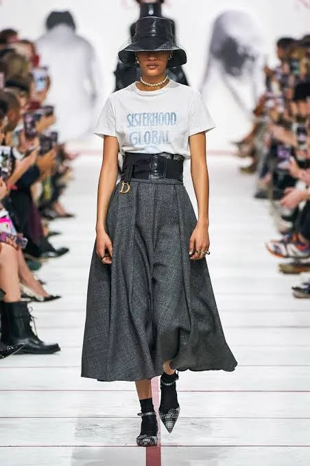 Runway model in a white SISTERHOOD GLOBAL t-shirt, grey midi skirt, black belt, and hat