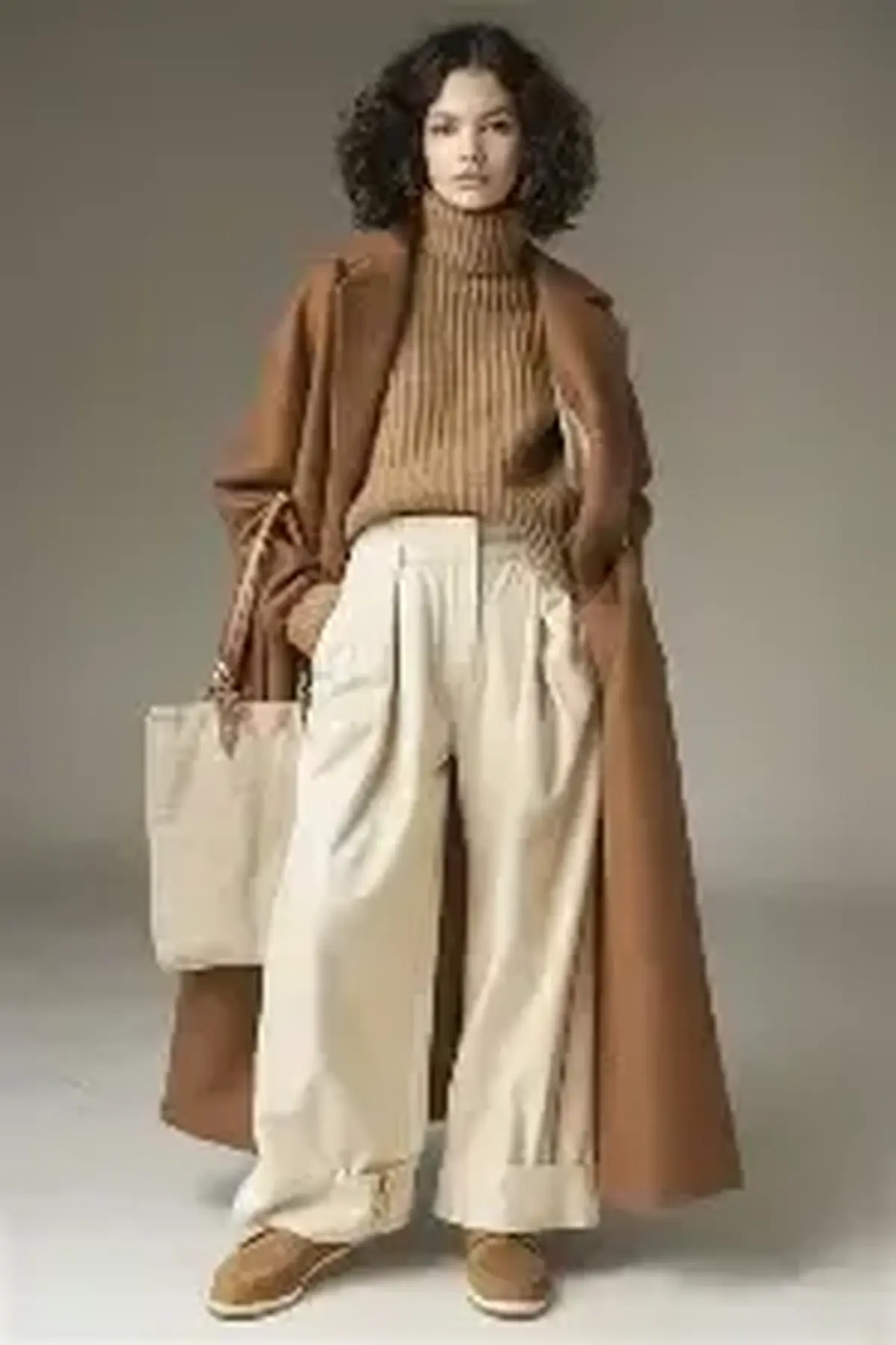 Woman in a long camel coat, camel ribbed turtleneck, cream wide-leg trousers, tan shoes and canvas tote against a grey backdrop