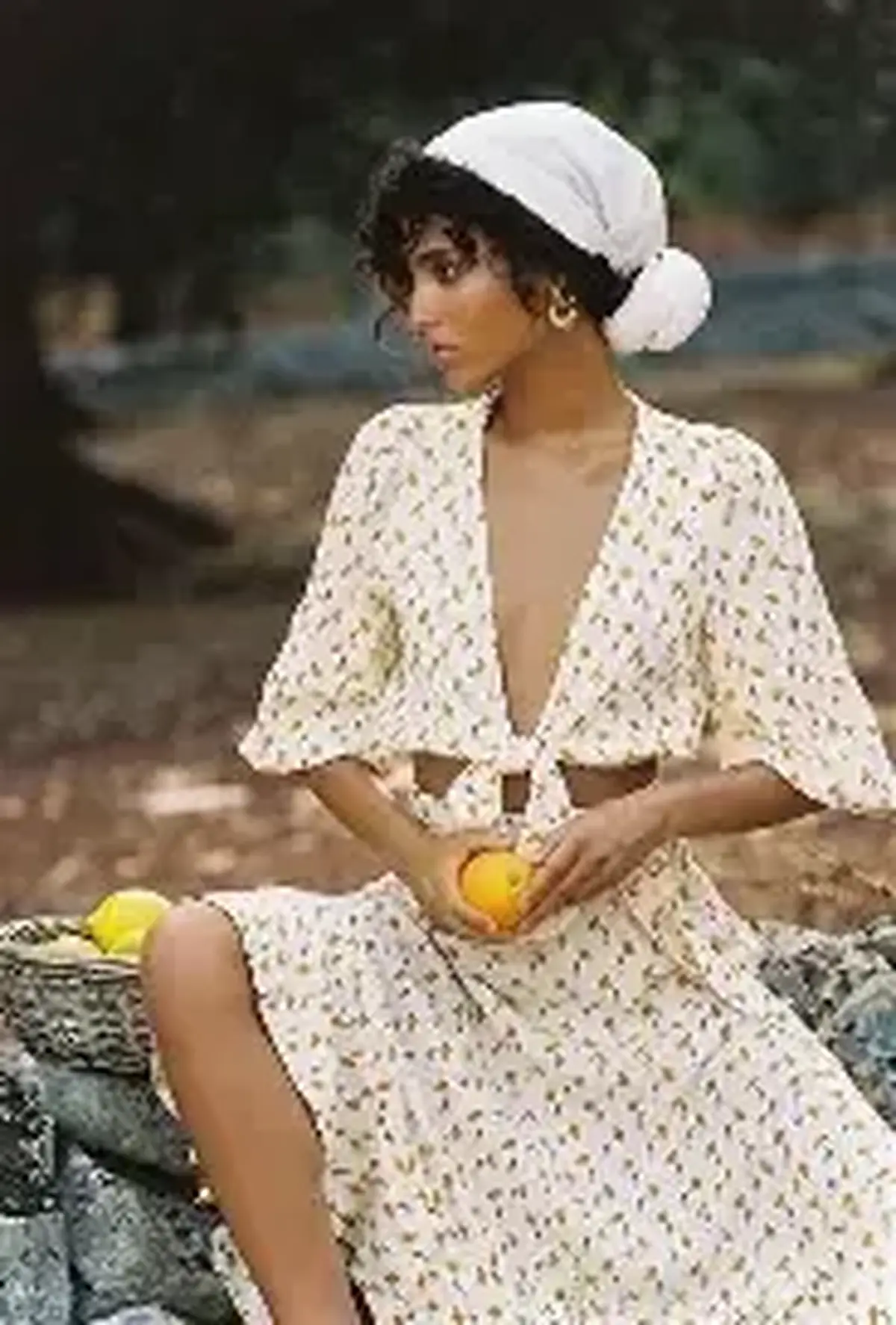 Woman in a white head-wrap and white floral tie-front outfit holding lemons in dappled shade