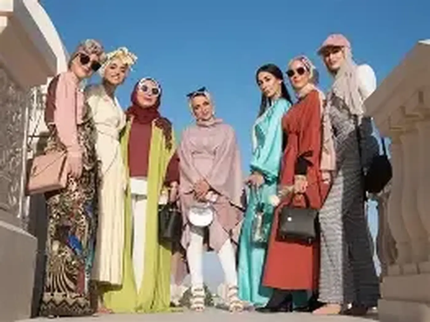 Group of women in pastel-toned hijabs, abayas, kaftans and long coats posing on a stone terrace under a blue sky