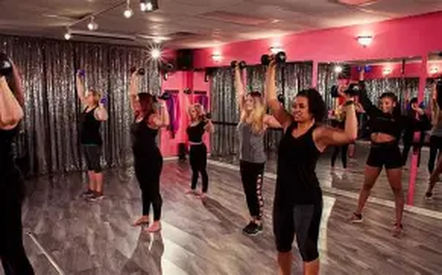 Women in black activewear lifting light dumbbells overhead in a pink mirrored studio