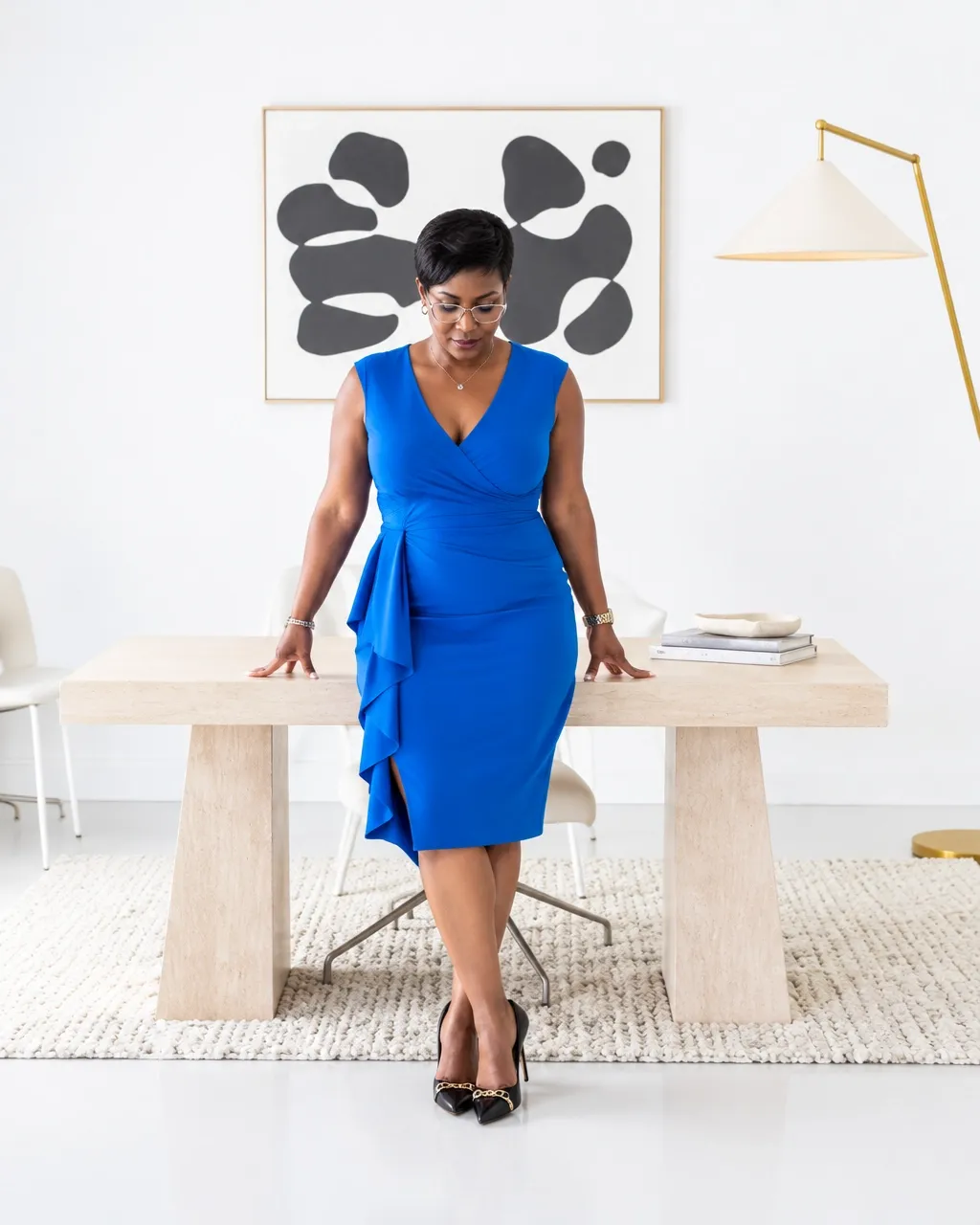 Natacha Pennycooke in a royal blue dress standing behind a pale wood desk with abstract artwork behind her