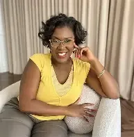 Natacha Pennycooke in a soft yellow top and glasses, laughing in a cream armchair