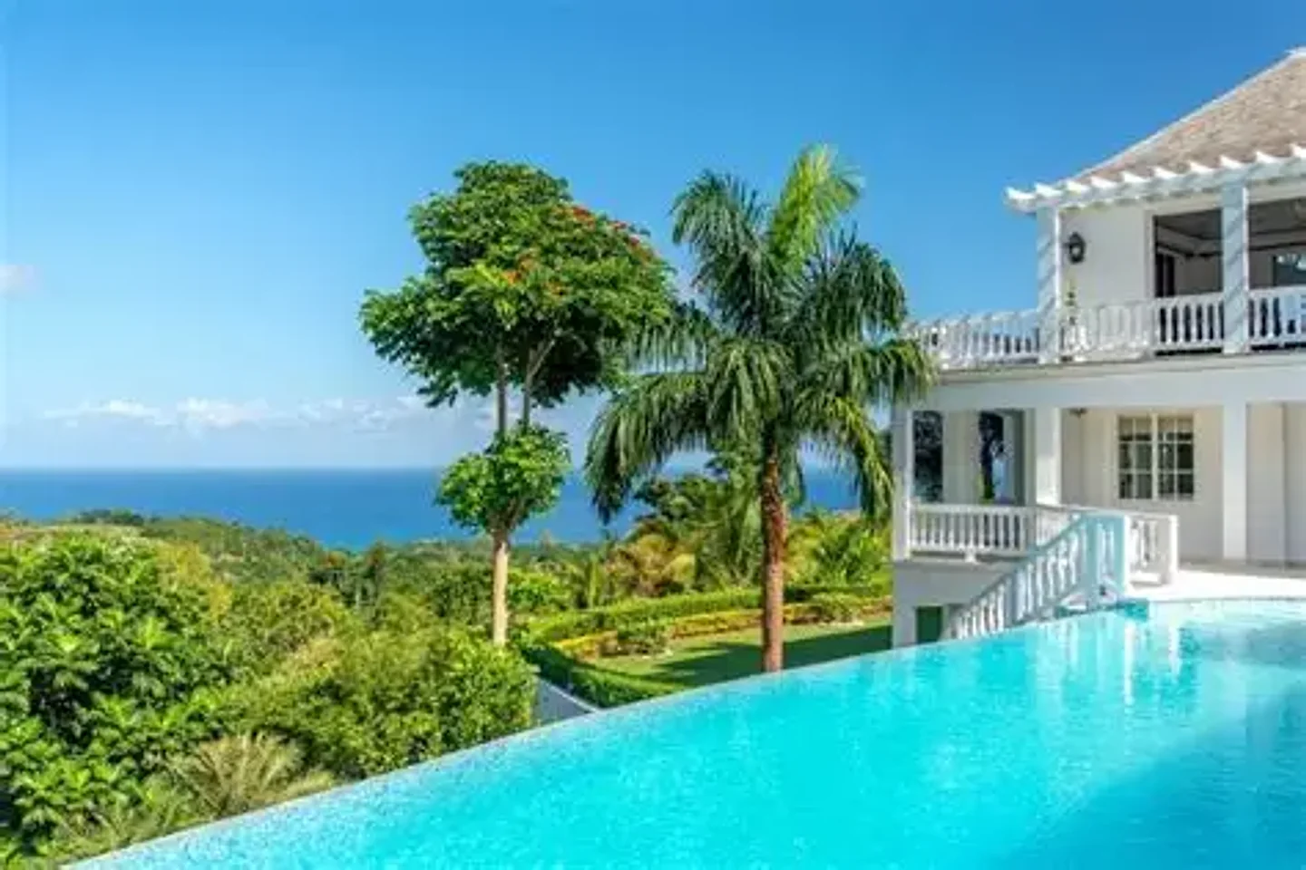 White villa with a veranda above a vivid turquoise infinity pool, palm trees and green hillside, ocean, and blue sky beyond