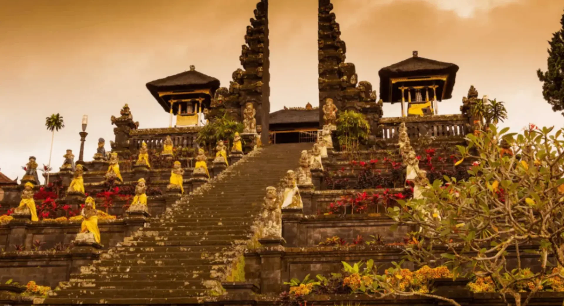 Tall stone staircase at a Balinese temple lined with statues wrapped in yellow and white cloth, red flowers, and warm sky