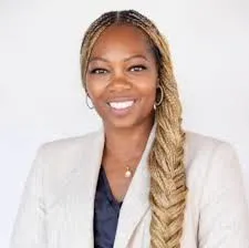 Portrait of Eronmwon Balogun smiling in a white blazer, with long braided hair, against a plain background