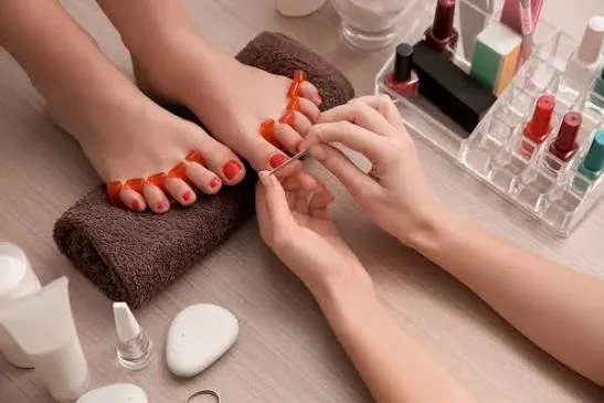 A woman receiving a pedicure, her feet resting on a rolled towel while a technician paints her toenails a warm coral shade