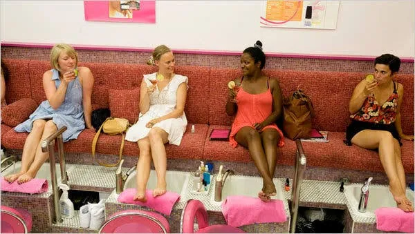A group of women relaxing together on a red couch at a nail salon, laughing and enjoying drinks during their pedicures