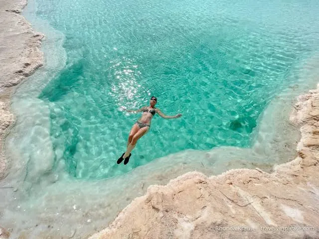 A woman floating weightless on her back in a turquoise mineral pool edged with pale stone