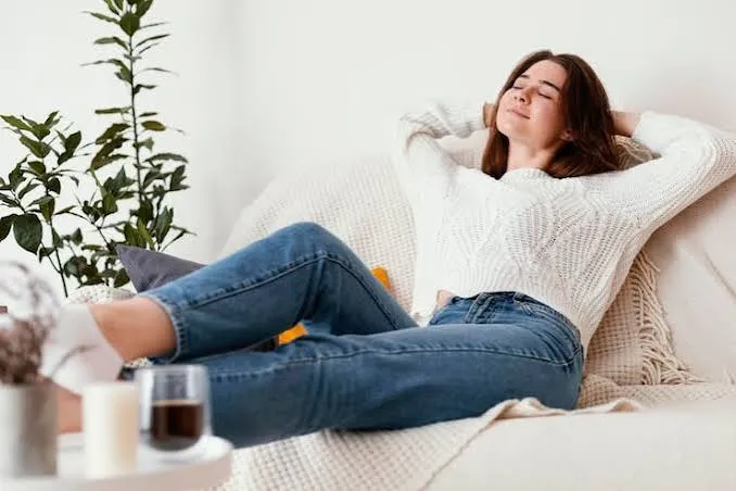 A woman in a white knit sweater leaning back with closed eyes on a cream couch, relaxed in soft morning light